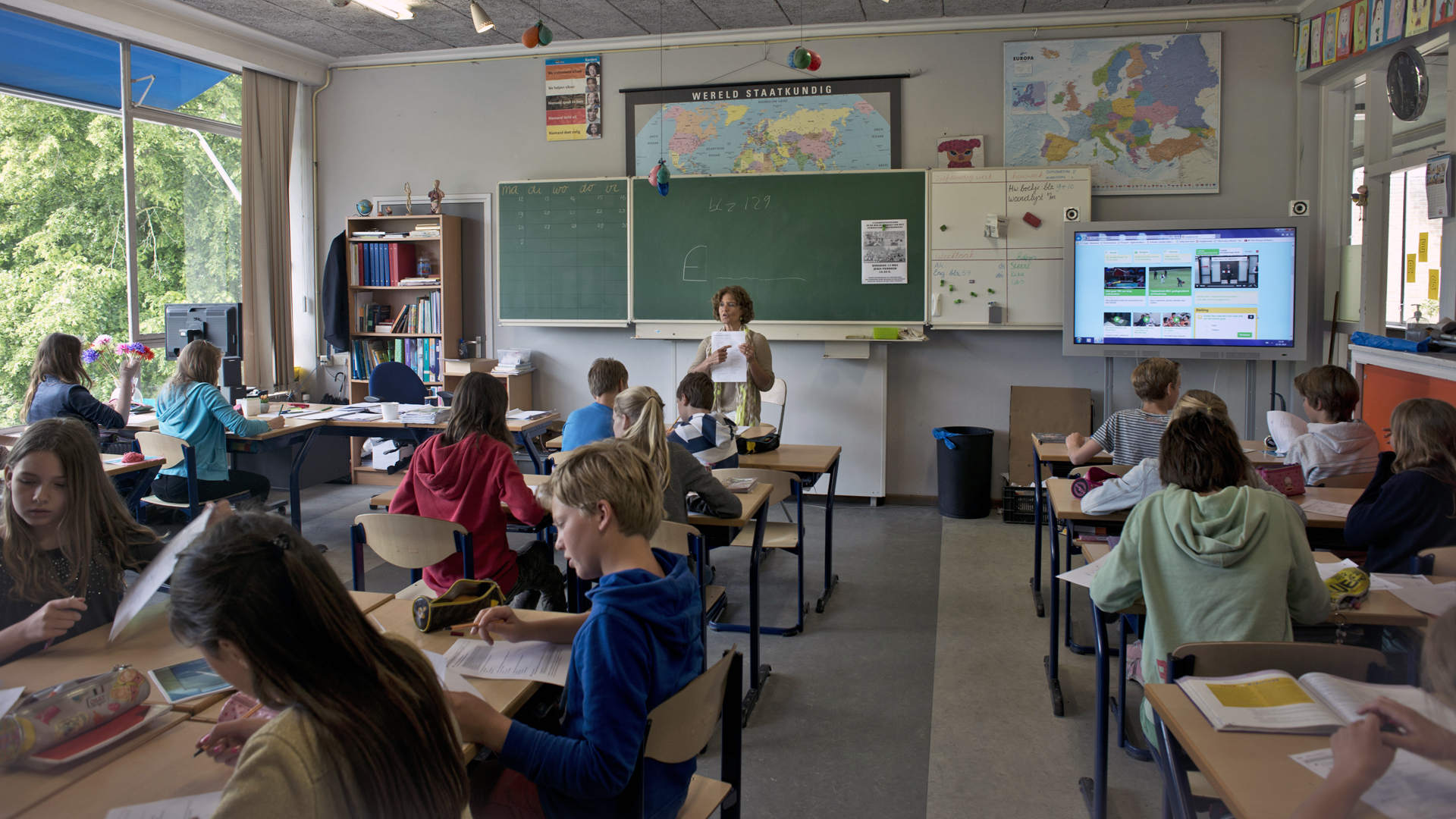 Een klaslokaal met kinderen aan tafels en een leerkracht voor de klas