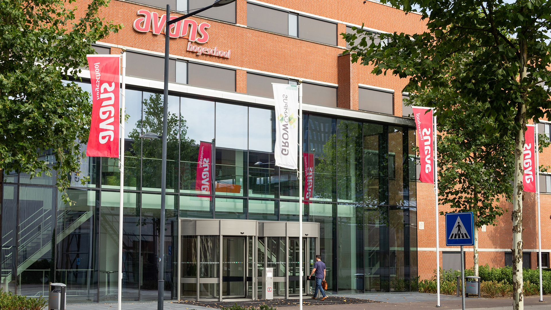 Voorkant van het Avans Hogeschool-gebouw aan de Onderwijsboulevard in Den Bosch, met een glazen entree, rode Avans-vlaggen en een persoon die naar binnen loopt.