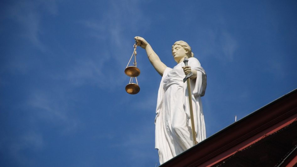 Standbeeld van Vrouwe Justitia met een zwaard en een weegschaal, gezien van onderaf tegen een heldere blauwe lucht