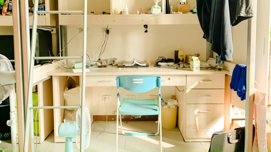 Rommelige studentenkamer met een wit bureau onder een hoogslaper, een lichtblauwe klapstoel ervoor, papieren, kabels en kleine voorwerpen op het bureau, kleding die aan de zijkant hangt en zonlicht dat door het raam naar binnen valt.