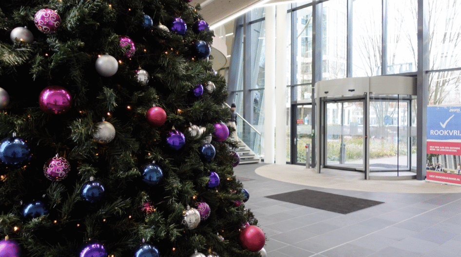 Een feestelijke kerstboom, versierd met roze, paarse en blauwe ornamenten, staat op de voorgrond van de moderne, lichte Avanslobby met grote glazen deuren aan de Hogeschoollaan.
